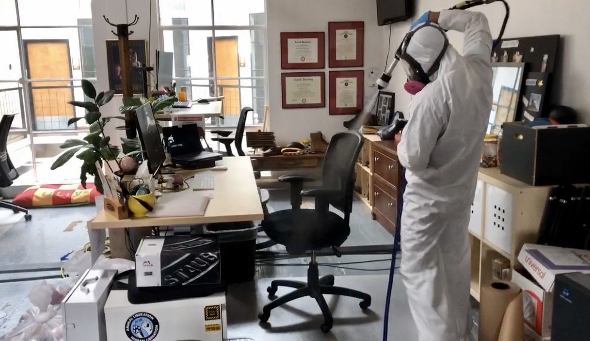 A Cleansolution employee wearing personal protective equipment uses an electrostatic sprayer to disinfect a black office chair.