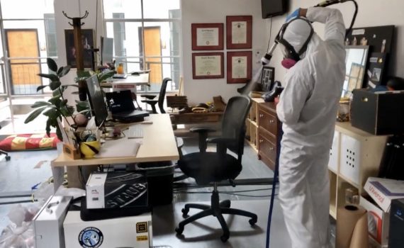 A Cleansolution employee wearing personal protective equipment uses an electrostatic sprayer to disinfect a black office chair.